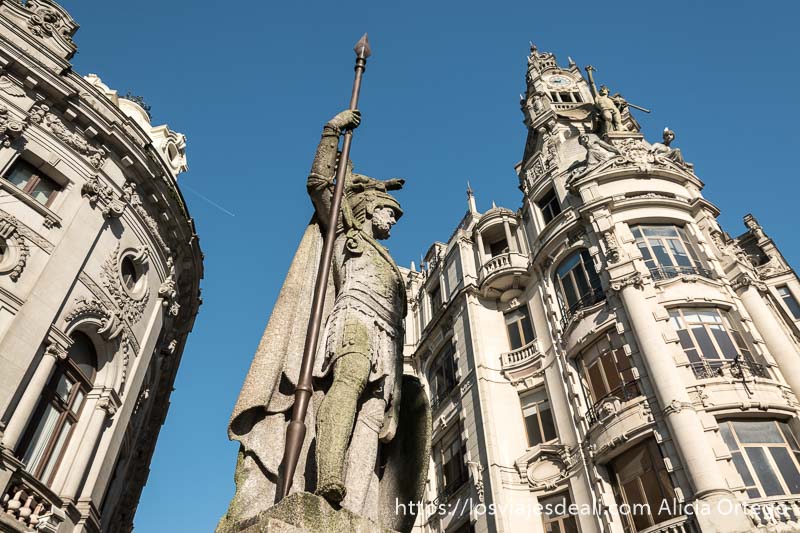 estatua de piedra de guerrero con casco apoyado en su lanza visto desde abajo y detrás edificios modernistas bajo cielo muy azul