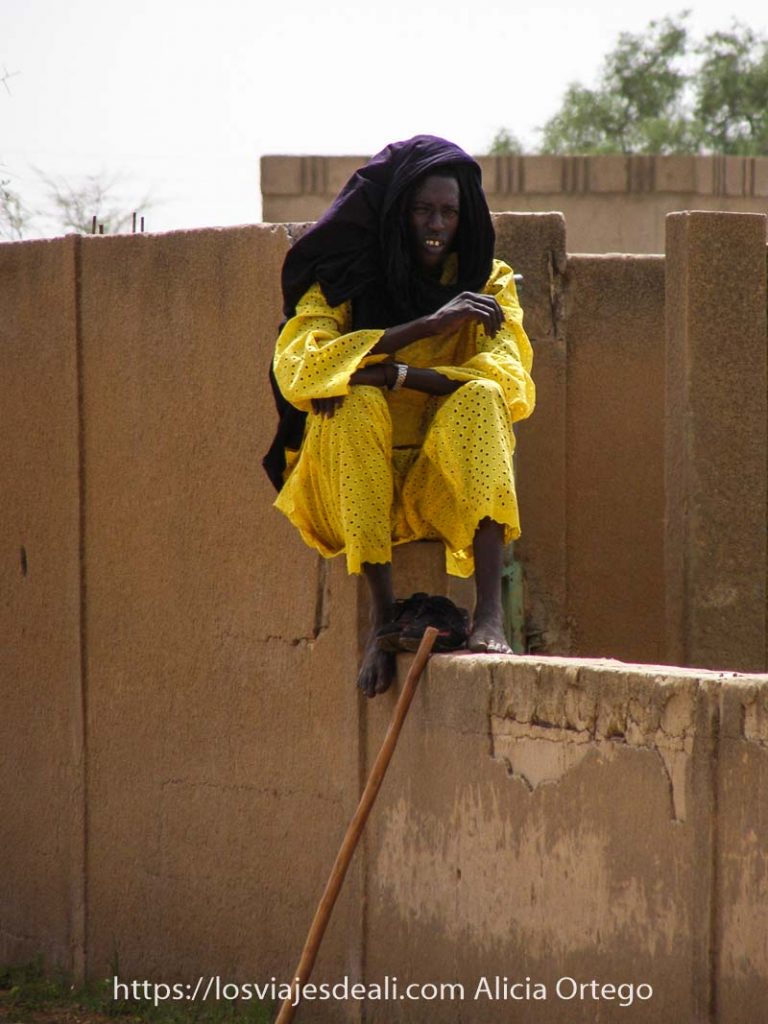 Mi primer mercado africano en el Sahel de Burkina Faso: Gorom Gorom 66 hombre muy delgado con traje amarillo chillón y turbante morado en la cabeza sentado en un muro de adobe en el mercado africano de gorom gorom